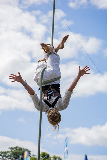 Areal performer suspended on rope in mid air. The performance is outside with trees in the background.