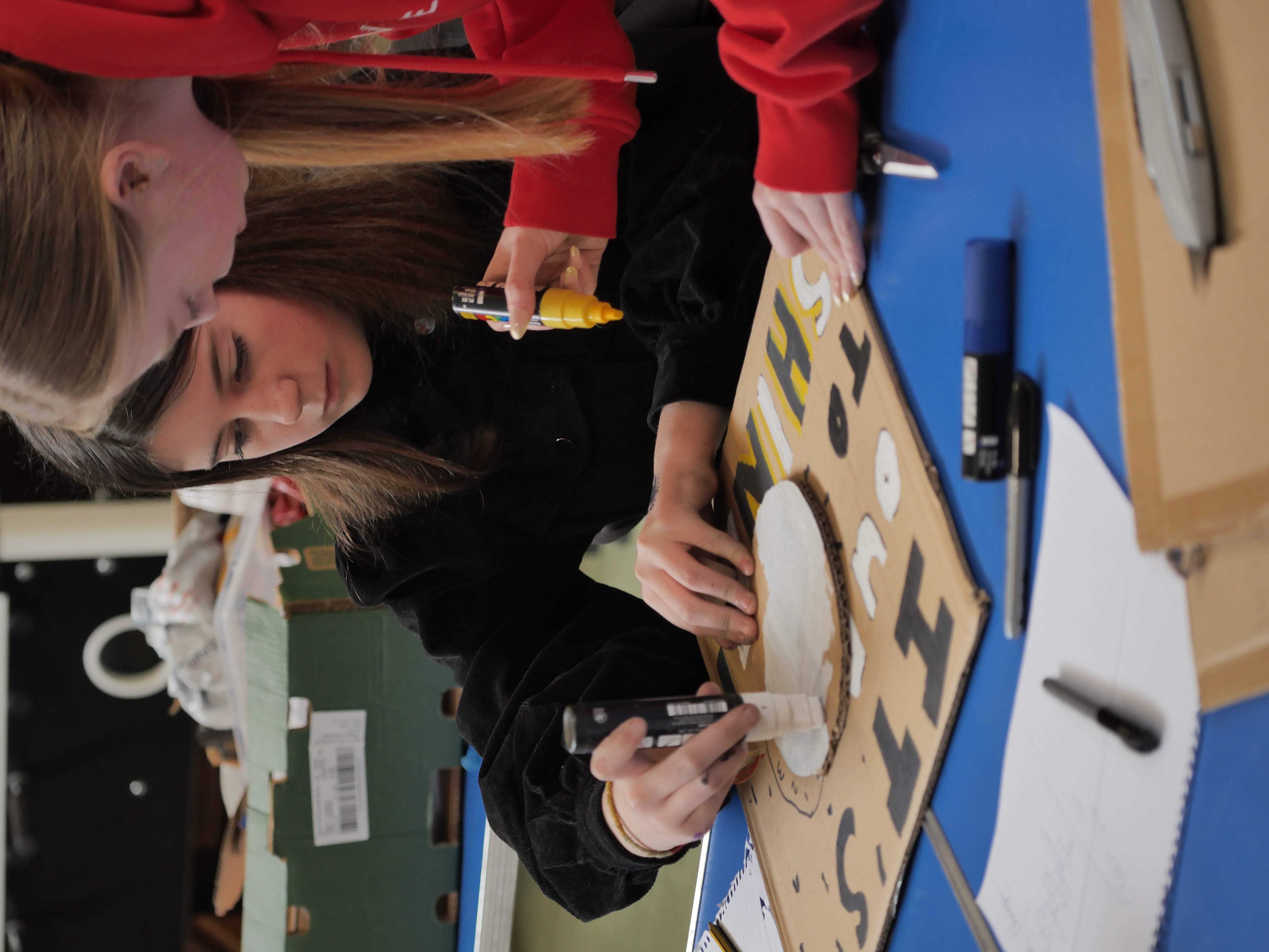 two young people working together on a craft project with pens and cardboard.