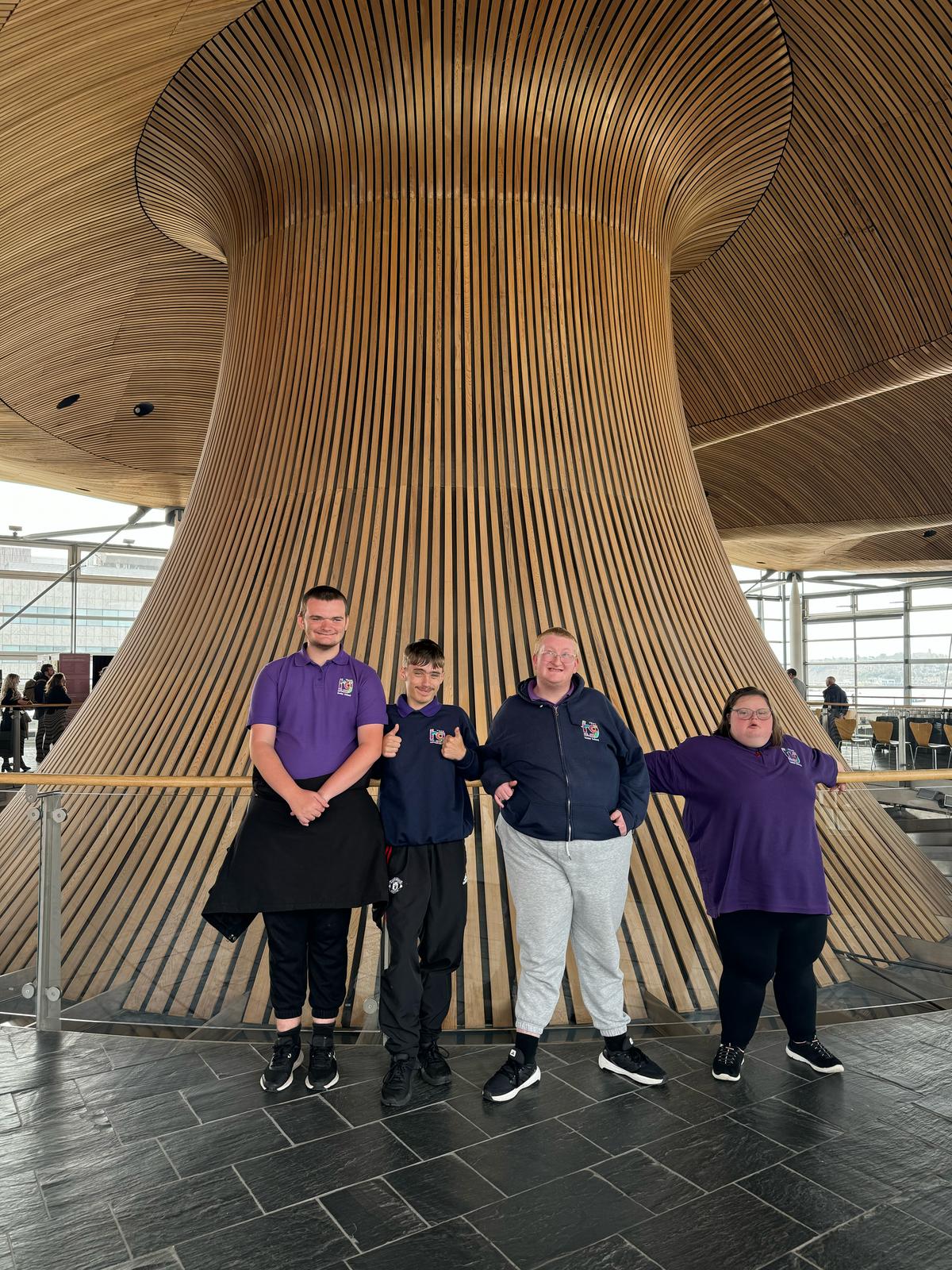 Four Volunteens standing in front of the iconic wooden funnel inside the Senedd building in Cardiff Bay during their visit to meet Jane Hutt MS